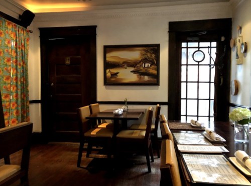 Cozy restaurant interior with wooden furniture, colorful curtains, and a landscape painting on the wall.