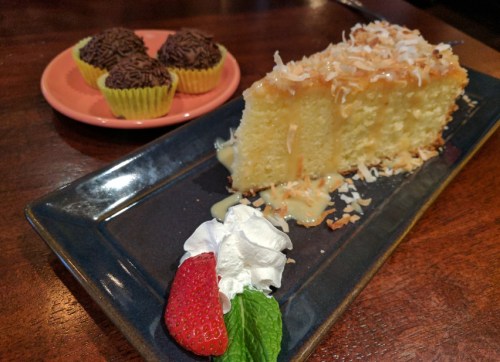 A slice of coconut cake drizzled with sauce, served with whipped cream, a strawberry, and a plate of chocolate-covered cupcakes.
