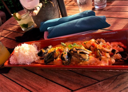 A vibrant dish of seafood pasta with mussels, served with rice, on a wooden table with drinks in the background.