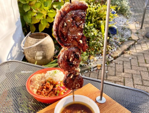 A skewer of grilled meat served with rice, beans, and a dipping sauce on a table outdoors.