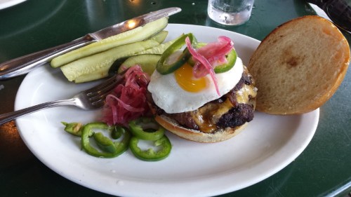 A juicy burger topped with a fried egg, jalapeños, and pickled onions, served with pickles on the side.
