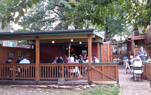 A cozy outdoor restaurant with wooden seating, patrons dining, and greenery surrounding the area.