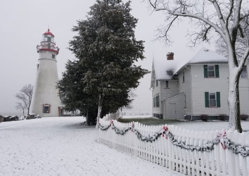 A snowy scene featuring a lighthouse and a house, with a white picket fence adorned with garlands.
