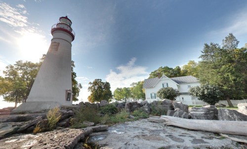 A tall lighthouse beside a white house, surrounded by trees and rocky shoreline under a clear blue sky.