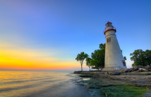 A lighthouse stands by the water at sunset, surrounded by trees and rocky shoreline under a colorful sky.