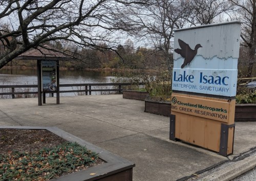 Sign for Lake Isaac Waterfowl Sanctuary near a calm lake, surrounded by trees and autumn foliage.