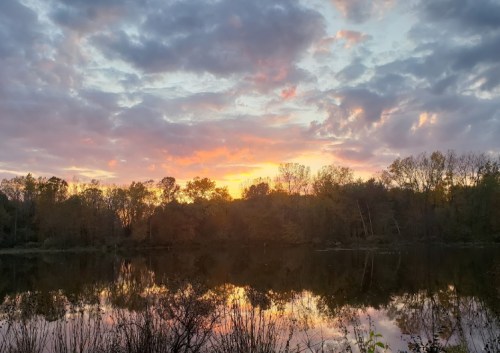 A serene sunset over a calm lake, with colorful clouds reflecting on the water and trees lining the shore.