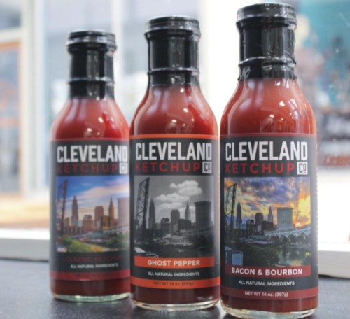 Three bottles of Cleveland Ketchup Co. featuring Classic, Ghost Pepper, and Bacon & Bourbon flavors against a city skyline.