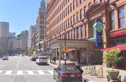 Street view of a city block featuring a Residence Inn, shops, and restaurants, with pedestrians and parked cars.