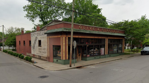 A brick building with "Olive & Sinclair" signage, featuring large windows and a tree-lined street.