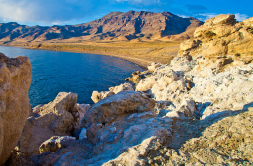 Rocky shoreline with clear blue water and mountains in the background under a bright sky.