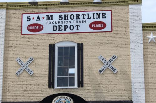 Sign for Sam Shortline Excursion Train Depot, featuring a large window and railroad crossing signs on a brick building.