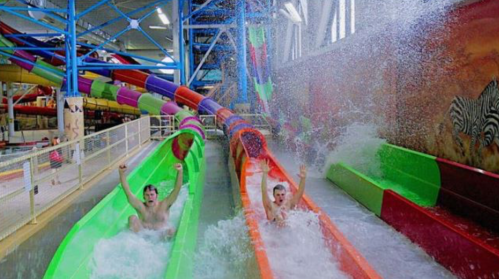 Two young men slide down colorful water slides, splashing into the pool below, with water spraying around them.