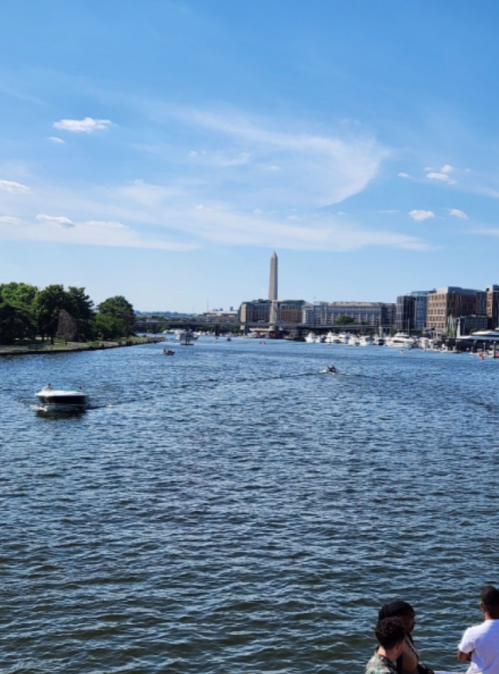 A river scene with boats, trees, and a tall monument in the background under a clear blue sky.
