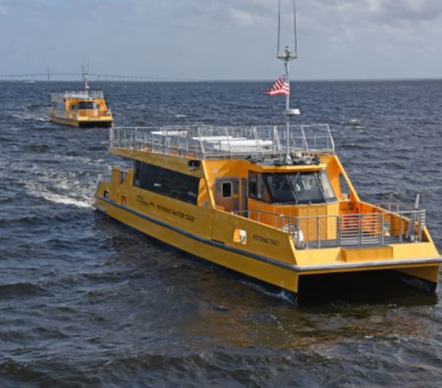 Two yellow boats navigate through water, with a bridge visible in the background and an American flag on one boat.