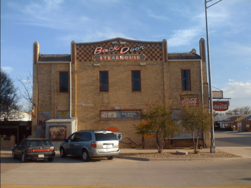 A brick building with a sign reading "Back Door Steakhouse," featuring a parking lot and nearby traffic signals.