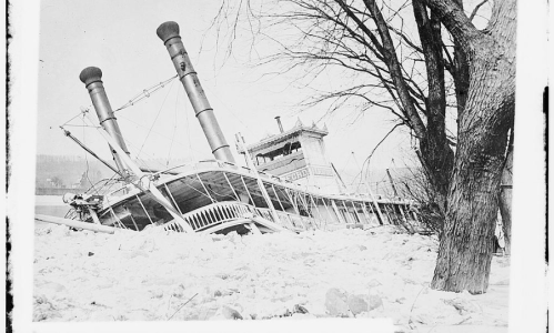 A steamboat partially submerged in ice, leaning against a tree in a winter landscape.