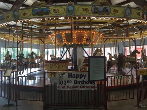 A colorful carousel with ornate horses, decorated for a birthday celebration, featuring a sign that reads "Happy 103rd Birthday."