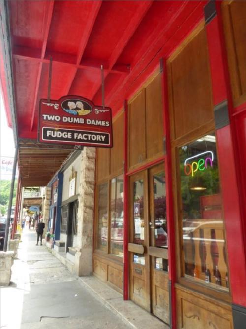 Sign for "Two Dumb Dames Fudge Factory" hanging above a storefront with an "open" neon sign.