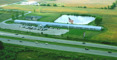 Aerial view of an antique shop with a large American flag, surrounded by fields and a pond, near a highway.
