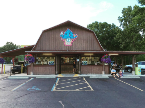 A wooden building with a colorful ice cream sign, featuring flower pots and people sitting outside.