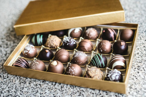 A gold box filled with assorted chocolate truffles, featuring various designs and toppings, on a speckled surface.