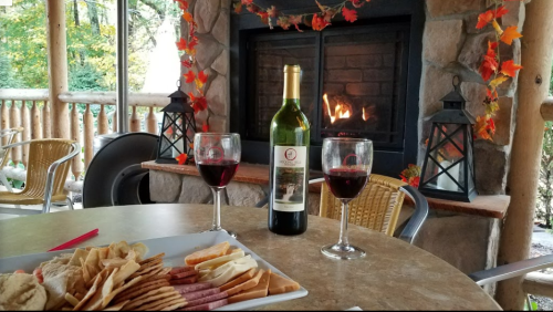 A cozy setting with a bottle of wine, two glasses, and a platter of snacks by a fireplace adorned with autumn leaves.