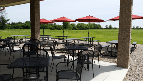 Outdoor seating area with black tables and chairs under red umbrellas, overlooking a green field.