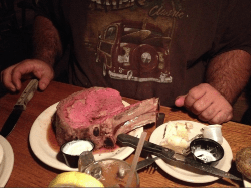 A large, juicy prime rib steak on a plate, accompanied by butter and dipping sauces, with a person’s hands nearby.