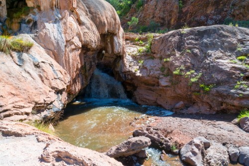 A rocky landscape with a small waterfall flowing into a pool, surrounded by rugged terrain and greenery.