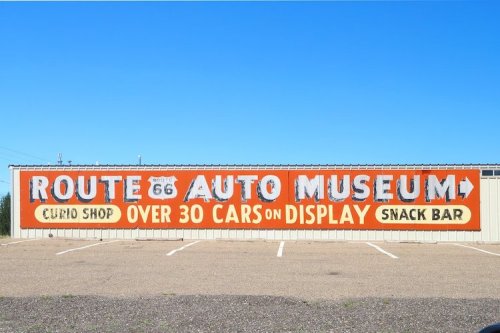 Colorful sign for Route 66 Auto Museum featuring "Curio Shop," "Over 30 Cars on Display," and "Snack Bar." Clear blue sky.
