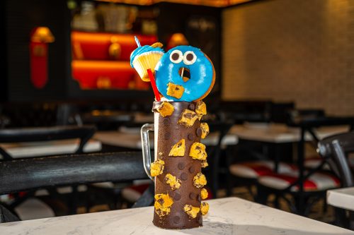 A colorful milkshake topped with a blue donut, cupcake, and cookie pieces, served in a tall glass on a table.