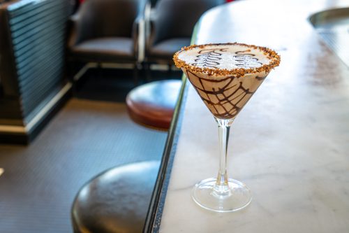 A chocolate martini with a decorative rim sits on a marble bar, with blurred seating in the background.