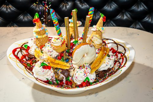 A large, colorful ice cream sundae topped with cones, sprinkles, cherries, and various sweets in a white bowl.