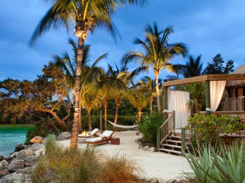 Tropical beach scene with palm trees, lounge chairs, and a cozy wooden cabin under a twilight sky.