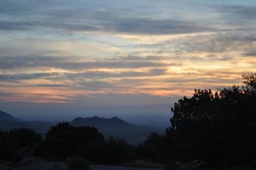 A serene sunset over distant mountains, with soft clouds and a colorful sky, framed by silhouettes of trees.