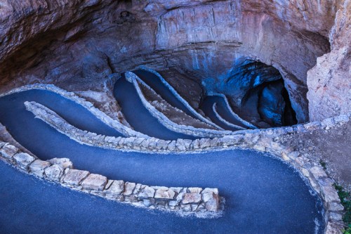 Winding stone path leading into a dark cave, surrounded by rocky walls and a textured, natural landscape.