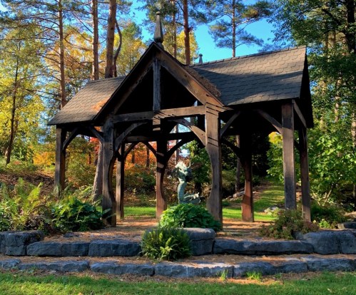 A wooden gazebo surrounded by greenery and trees, with a stone seating area in a serene outdoor setting.