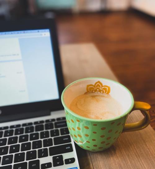 A green polka-dotted coffee mug sits next to a laptop on a wooden table.