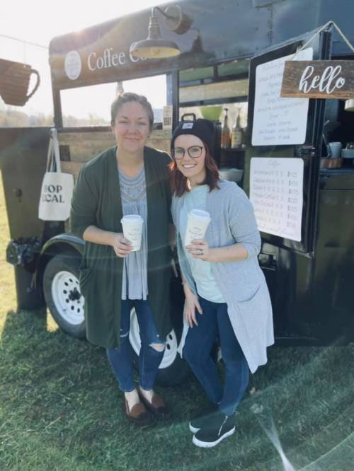 Two women stand in front of a coffee truck, each holding a coffee cup, smiling in a sunny outdoor setting.