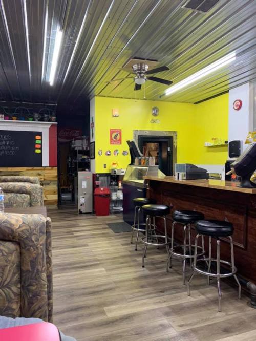 A brightly lit café with yellow walls, a wooden counter, bar stools, and a cozy seating area.