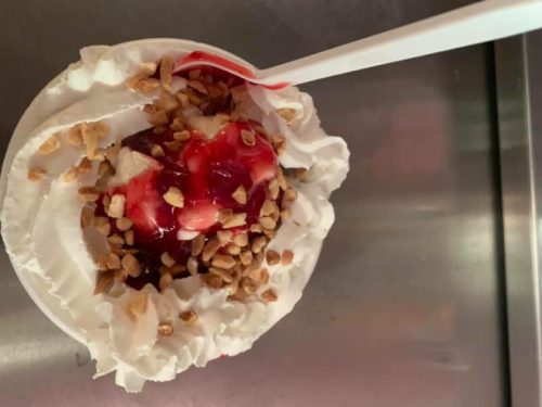 A dessert cup topped with whipped cream, cherry sauce, and chopped nuts, served with a plastic spoon.