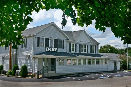 A white, two-story building with "Log Inn" sign, surrounded by greenery and a clear blue sky. Parking area in front.
