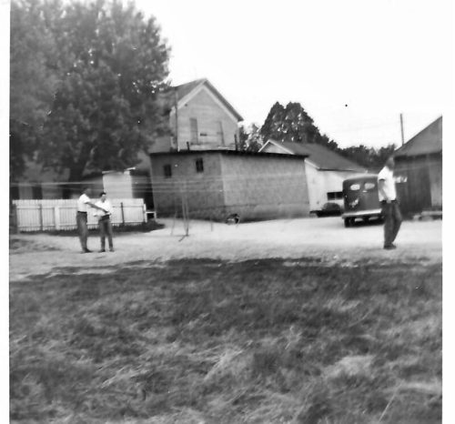Two men stand in a gravel area near a vintage car, with houses and trees in the background. Black and white photo.