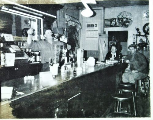A vintage bar scene with a bartender serving drinks, two patrons seated at the counter, and retro decor in the background.
