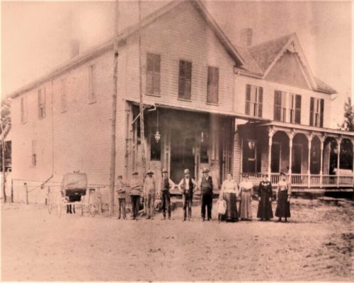 A historic black-and-white photo of a group of people standing in front of a large house with a porch and a horse-drawn carriage.