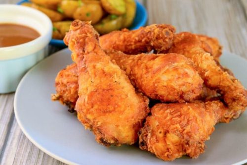 A plate of crispy fried chicken drumsticks with a side of dipping sauce and golden-brown potato wedges.