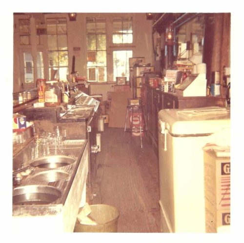 A vintage diner interior with a counter, sinks, and various kitchen items, illuminated by natural light from windows.