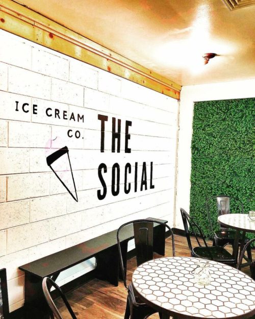 Interior of an ice cream shop featuring a wall with "THE SOCIAL" sign and green foliage decor. Tables and chairs are set up.