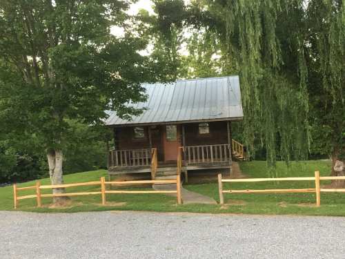 A rustic wooden cabin with a metal roof, surrounded by trees and a grassy area, with a gravel path and wooden fence.